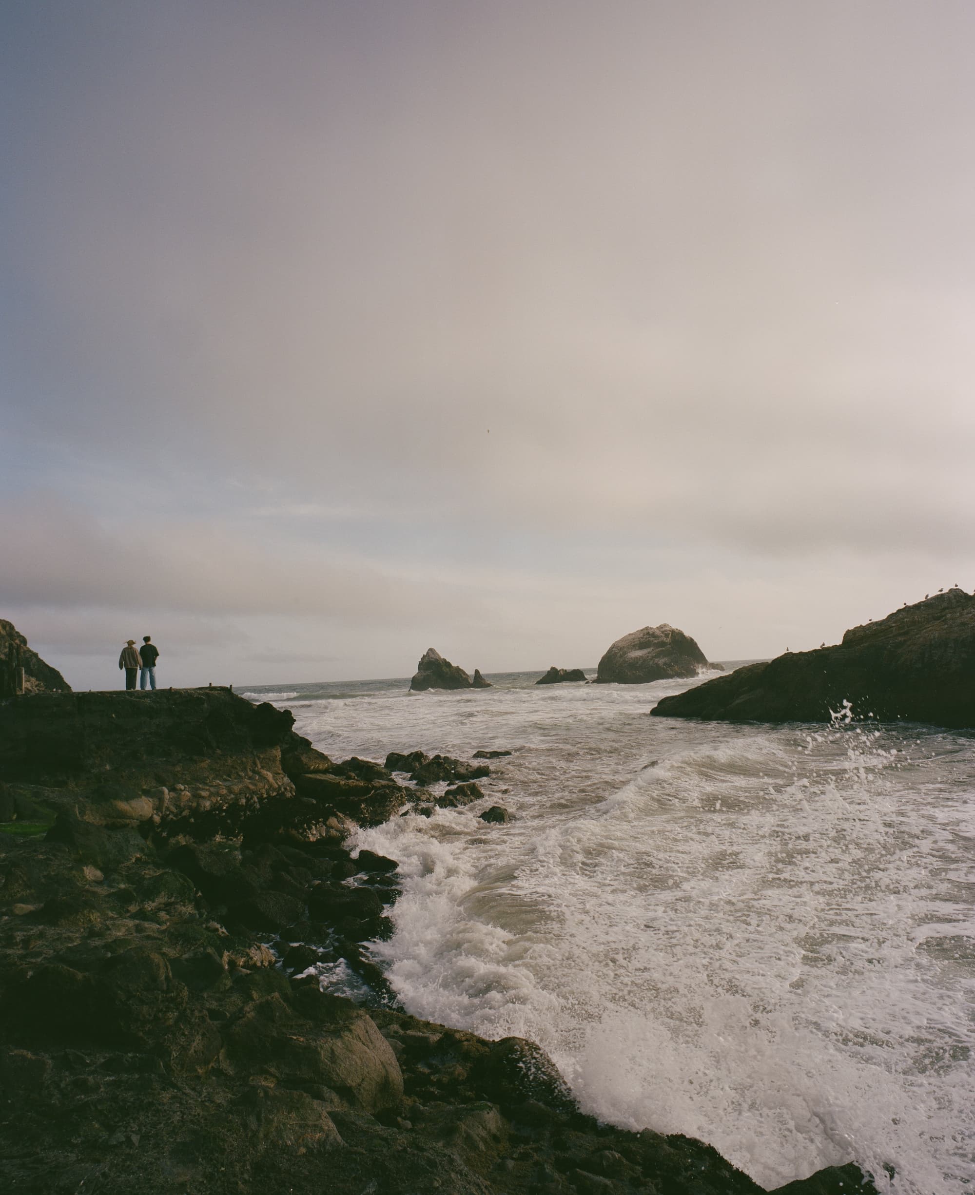 Film photograph — two figures at the boundary where land meets sea